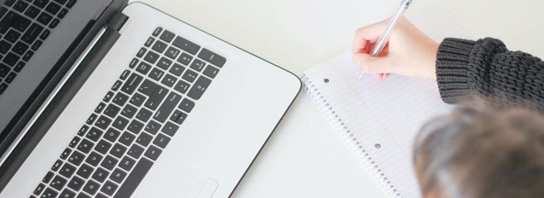 Laptop on a desk with a woman writing on a paper notepad next to it.