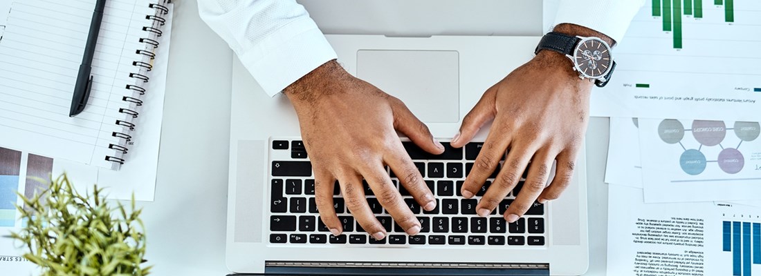 Person typing on a laptop with documents, graphs, and a notebook on the desk