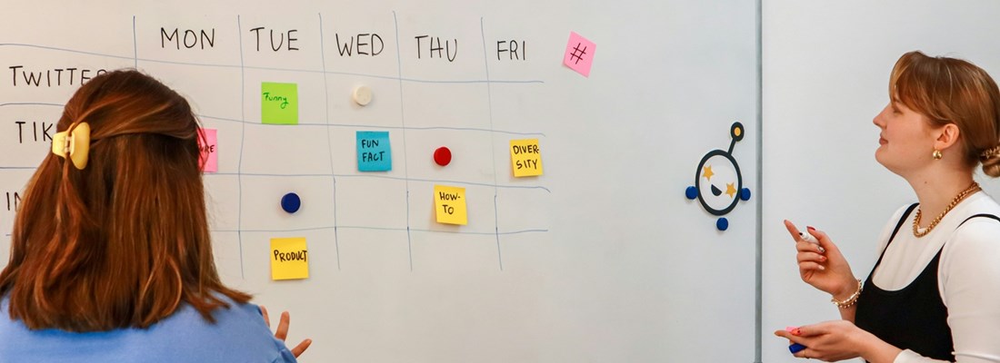 Two professionals planning a weekly social media calendar for accounting content on a whiteboard, with platforms like LinkedIn, TikTok, and Twitter