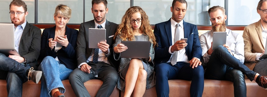 Group of business professionals using laptops, tablets, and smartphones in a modern office with large windows