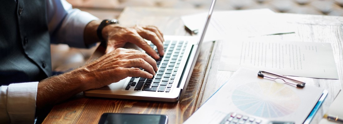 Person typing on a laptop at a wooden desk with financial documents, calculator, and smartphone